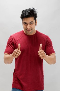 Mutlu, kendine güveni olan kırmızı tişörtlü bir adam, çifte baş parmak işareti yapıyor, başarıyı, pozitifliği, onayı ve cesaretlendirmeyi simgeliyor. Beyaz arkaplanda stüdyo resmi.