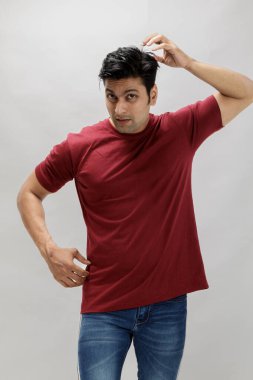 Kırmızı tişörtlü, kafası karışık bir ifadeyle kaşınan açık gri arka planda duran genç bir adamın portresi. Şüphe kavramı, problem çözme ve günlük yaşam tarzı..