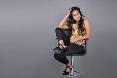 Çiçekli bir bluz giyen genç bir kadın, siyah taytlar ve siyah topuklular, sandalye ve neşeli yüz ifadeleri ile poz veriyor ve modern tarzı, zarafeti gösteren farklı duyguları var..