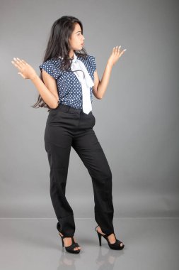 İş kıyafetleri içinde, siyah pantolon ve siyah topuklu ayakkabı giyen, sade bir arka plana karşı poz veren kendinden emin genç bir kadın.