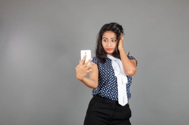 Kendine güveni tam bir genç kadın iş kıyafetleri içinde, puantiyeli bluz, siyah pantolon, siyah topuklu ayakkabı giyiyor, selfie için poz veriyor, eğlenceli ve hareketli bir jest yapıyor, oyunculuğu vurguluyor..