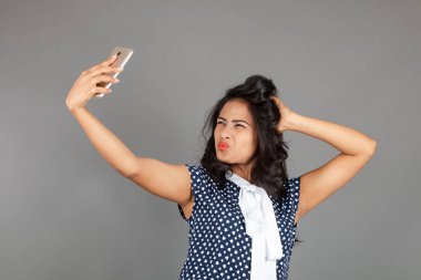 Kendine güveni tam bir genç kadın iş kıyafetleri içinde, puantiyeli bluz, siyah pantolon, siyah topuklu ayakkabı giyiyor, selfie için poz veriyor, eğlenceli ve hareketli bir jest yapıyor, oyunculuğu vurguluyor..