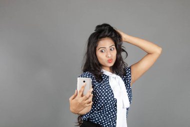 Kendine güveni tam bir genç kadın iş kıyafetleri içinde, puantiyeli bluz, siyah pantolon, siyah topuklu ayakkabı giyiyor, selfie için poz veriyor, eğlenceli ve hareketli bir jest yapıyor, oyunculuğu vurguluyor..