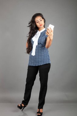 Kendine güveni tam bir genç kadın iş kıyafetleri içinde, puantiyeli bluz, siyah pantolon, siyah topuklu ayakkabı giyiyor, selfie için poz veriyor, eğlenceli ve hareketli bir jest yapıyor, oyunculuğu vurguluyor..