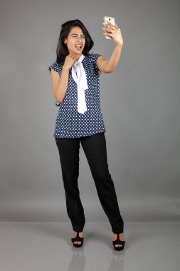 Kendine güveni tam bir genç kadın iş kıyafetleri içinde, puantiyeli bluz, siyah pantolon, siyah topuklu ayakkabı giyiyor, selfie için poz veriyor, eğlenceli ve hareketli bir jest yapıyor, oyunculuğu vurguluyor..