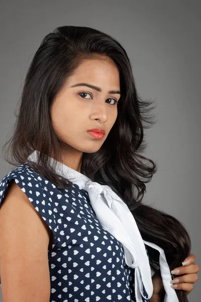 Kendine güvenen, şık bir genç kadın, puantiyeli bluz ve siyah pantolon giyiyor, sade bir arka plana karşı poz veriyor.