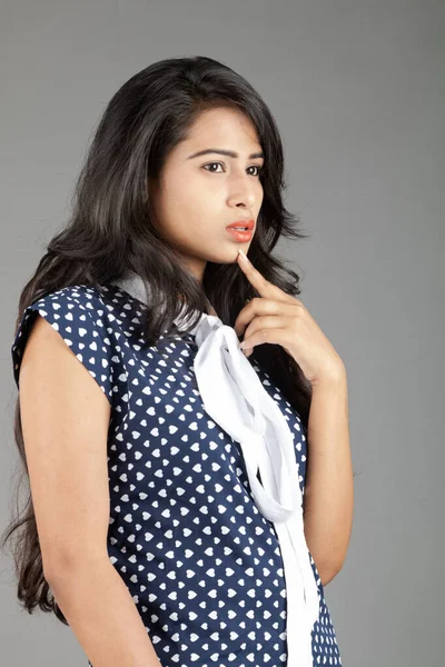 Kendine güvenen, şık bir genç kadın, puantiyeli bluz ve siyah pantolon giyiyor, sade bir arka plana karşı poz veriyor.