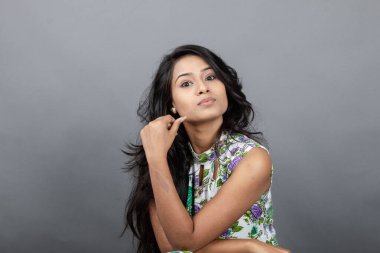 Kendine güvenen, çiçekli bluz giyen genç bir kadın zarif bir şekilde stüdyo ortamında poz veriyor. Güzelliğini, kendine güvenini ve tarzını sergiliyor. Onun uzun saçları zarif bir şekilde akıyor, resme hareketlilik ve canlılık katıyor.