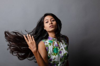 Kendine güvenen, çiçekli bluz giyen genç bir kadın zarif bir şekilde stüdyo ortamında poz veriyor. Güzelliğini, kendine güvenini ve tarzını sergiliyor. Onun uzun saçları zarif bir şekilde akıyor, resme hareketlilik ve canlılık katıyor.