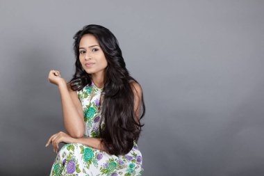 Kendine güvenen, çiçekli bluz giyen genç bir kadın zarif bir şekilde stüdyo ortamında poz veriyor. Güzelliğini, kendine güvenini ve tarzını sergiliyor. Onun uzun saçları zarif bir şekilde akıyor, resme hareketlilik ve canlılık katıyor.