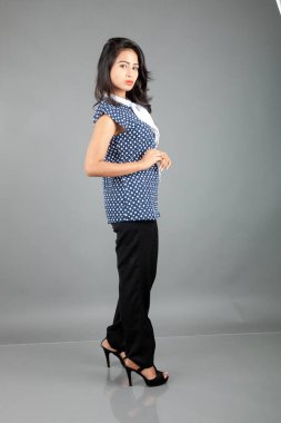 İş kıyafetleri içinde, siyah pantolon ve siyah topuklu ayakkabı giyen, sade bir arka plana karşı poz veren kendinden emin genç bir kadın.