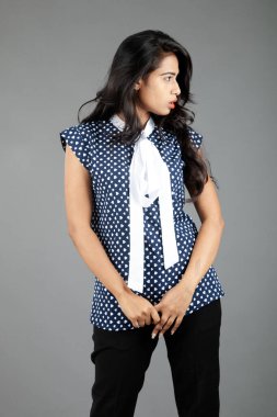 İş kıyafetleri içinde, siyah pantolon ve siyah topuklu ayakkabı giyen, sade bir arka plana karşı poz veren kendinden emin genç bir kadın.