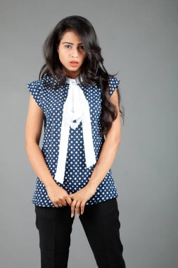 İş kıyafetleri içinde, siyah pantolon ve siyah topuklu ayakkabı giyen, sade bir arka plana karşı poz veren kendinden emin genç bir kadın.