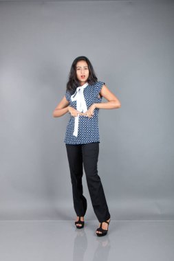 İş kıyafetleri içinde, siyah pantolon ve siyah topuklu ayakkabı giyen, sade bir arka plana karşı poz veren kendinden emin genç bir kadın.