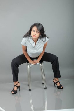 Gündelik iş kıyafetleri giymiş kendine güvenen bir kadın stüdyo ortamında modern bir taburede oturuyor, profesyonelliği, zarafeti ve moda veya iş temalı projeler için uygun duruşu çağrıştırıyor..