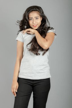 Gri polo tişörtlü, siyah pantolonlu, siyah topuklu neşeli genç bir kadın. Stüdyoda gri bir arka planda dalgalı saçla poz veriyor. Neşeli bir ifadesi var..