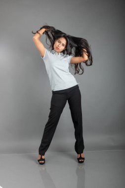 Gri polo tişörtlü, siyah pantolonlu, siyah topuklu neşeli genç bir kadın. Stüdyoda gri bir arka planda dalgalı saçla poz veriyor. Neşeli bir ifadesi var..