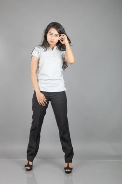 Gri polo tişörtlü, siyah pantolonlu, siyah topuklu neşeli genç bir kadın. Stüdyoda gri bir arka planda dalgalı saçla poz veriyor. Neşeli bir ifadesi var..