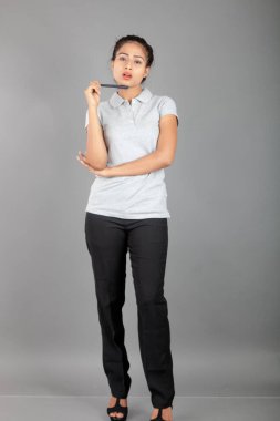 Neşeli bir ofis hanımı ya da kalemle poz veren genç bir üniversite öğrencisi. Yarı resmi giyinmiş genç bir kadın gri polo tişört giyiyor, siyah pantolon ve gri arka plan..