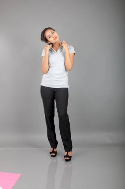 Gri polo gömlekli, siyah pantolonlu, siyah topuklu genç bir ofis çalışanı veya genç bir üniversite öğrencisi, gri bir arka planla stüdyo ortamında poz veriyor. Neşeli bir ifadesi var..