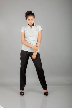 Gri polo gömlekli, siyah pantolonlu, siyah topuklu genç bir ofis çalışanı veya genç bir üniversite öğrencisi, gri bir arka planla stüdyo ortamında poz veriyor. Neşeli bir ifadesi var..