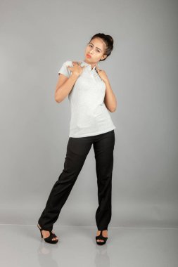 Gri polo gömlekli, siyah pantolonlu, siyah topuklu genç bir ofis çalışanı veya genç bir üniversite öğrencisi, gri bir arka planla stüdyo ortamında poz veriyor. Neşeli bir ifadesi var..