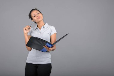 Neşeli ofis çalışanı ya da elinde bir dosya, kalem ve sunum tutan genç bir üniversite öğrencisi. Resmi giyinmiş genç bir kadın gri polo tişört giyiyor, siyah pantolon ve gri arka plan..