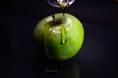 Koyu arkaplanda taze yeşil bir Granny Smith elmasına bal damlatan bir kadın eli.