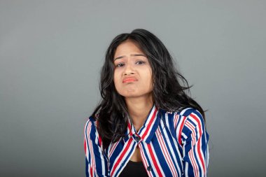 Üzgün Hintli genç kadın portresi somurtuyor, somurtuyor ve üzgün görünüyor, üzgün yüz ifadesi, gri arka plana karşı kasvetli duruyor.