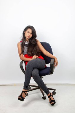Neşeli, genç, güzel bir Hintli kadın manken. Kırmızı ve gri tasarımcı bluzu ve siyah kot pantolon giyiyor. Beyaz arka planda bir sandalyede oturuyor. Moda Portresi. Reklam çekimi.