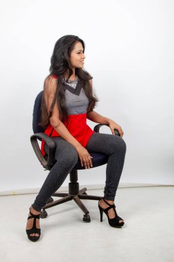 Neşeli, genç, güzel bir Hintli kadın manken. Kırmızı ve gri tasarımcı bluzu ve siyah kot pantolon giyiyor. Beyaz arka planda bir sandalyede oturuyor. Moda Portresi. Reklam çekimi.
