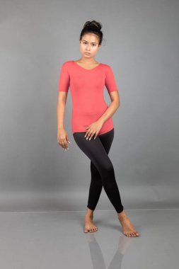 Kırmızı tişörtlü, siyah taytlı, yoga ve esneme egzersizleri yapan sağlıklı genç bir kadın gri arka planda izole edilmiş.