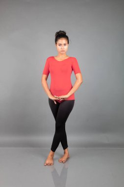 Kırmızı tişörtlü, siyah taytlı, yoga ve esneme egzersizleri yapan sağlıklı genç bir kadın gri arka planda izole edilmiş.