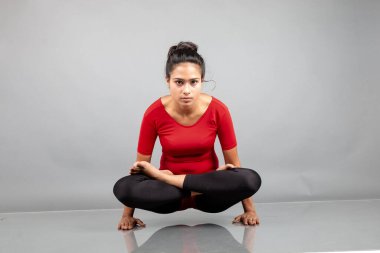 Kırmızı tişörtlü, siyah taytlı, yoga ve esneme egzersizleri yapan sağlıklı genç bir kadın gri arka planda izole edilmiş.