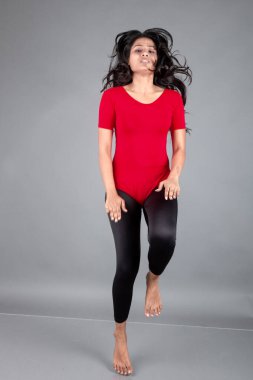 Kırmızı tişörtlü, siyah taytlı, yoga ve esneme egzersizleri yapan sağlıklı genç bir kadın gri arka planda izole edilmiş.