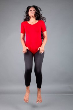 Kırmızı tişörtlü, siyah taytlı, yoga ve esneme egzersizleri yapan sağlıklı genç bir kadın gri arka planda izole edilmiş.