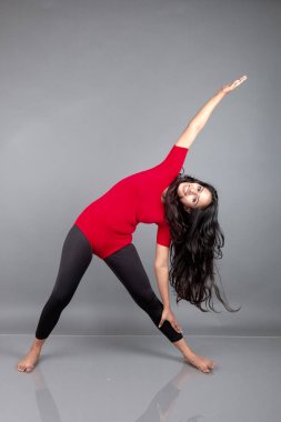 Kırmızı tişörtlü, siyah taytlı, yoga ve esneme egzersizleri yapan sağlıklı genç bir kadın gri arka planda izole edilmiş.