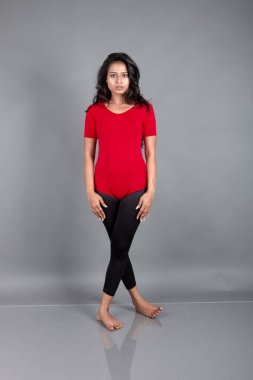 Kırmızı tişörtlü, siyah taytlı, yoga ve esneme egzersizleri yapan sağlıklı genç bir kadın gri arka planda izole edilmiş.