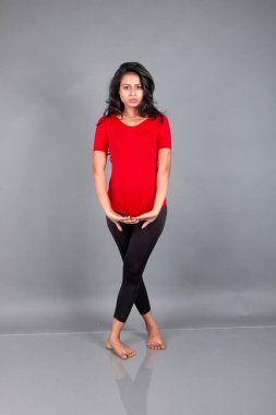 Kırmızı tişörtlü, siyah taytlı, yoga ve esneme egzersizleri yapan sağlıklı genç bir kadın gri arka planda izole edilmiş.