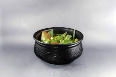 Yeşil dolma biber, kırmızı dolma biber, bebek mısırı, kiraz domatesi, kaffir limon yaprağı gibi sebzeli yeşil Hint cevizi tabanlı köri, beyaz arka planda siyah seramik bir kasede servis ediliyor.