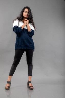 Gri arka planda günlük kışlık giysili Hintli genç bayan model. Siyah tayt giyen uzun siyah saçlı bir manken, mavi beyaz bir kazak..