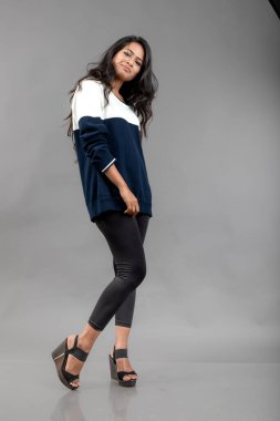 Gri arka planda günlük kışlık giysili Hintli genç bayan model. Siyah tayt giyen uzun siyah saçlı bir manken, mavi beyaz bir kazak..