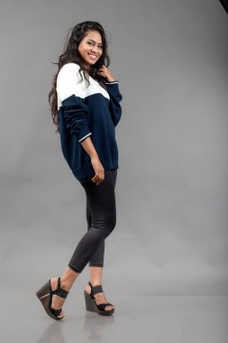 Gri arka planda günlük kışlık giysili Hintli genç bayan model. Siyah tayt giyen uzun siyah saçlı bir manken, mavi beyaz bir kazak..