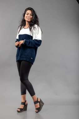 Gri arka planda günlük kışlık giysili Hintli genç bayan model. Siyah tayt giyen uzun siyah saçlı bir manken, mavi beyaz bir kazak..