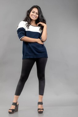 Gri arka planda günlük kışlık giysili Hintli genç bayan model. Siyah tayt giyen uzun siyah saçlı bir manken, mavi beyaz bir kazak..