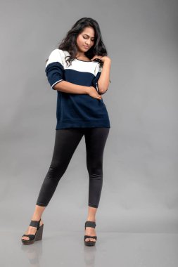 Gri arka planda günlük kışlık giysili Hintli genç bayan model. Siyah tayt giyen uzun siyah saçlı bir manken, mavi beyaz bir kazak..