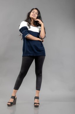 Gri arka planda günlük kışlık giysili Hintli genç bayan model. Siyah tayt giyen uzun siyah saçlı bir manken, mavi beyaz bir kazak..