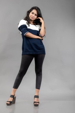 Gri arka planda günlük kışlık giysili Hintli genç bayan model. Siyah tayt giyen uzun siyah saçlı bir manken, mavi beyaz bir kazak..