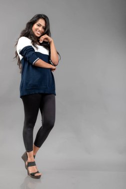 Gri arka planda günlük kışlık giysili Hintli genç bayan model. Siyah tayt giyen uzun siyah saçlı bir manken, mavi beyaz bir kazak..