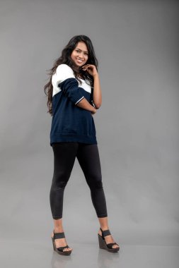 Gri arka planda günlük kışlık giysili Hintli genç bayan model. Siyah tayt giyen uzun siyah saçlı bir manken, mavi beyaz bir kazak..